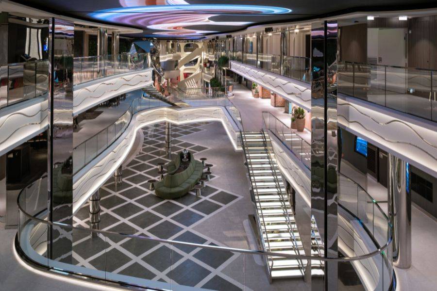 Modernes Atrium eines Kreuzfahrtschiffs mit mehreren Etagen, geschwungenen Glasgeländern, stilvollen Beleuchtungselementen und einem zentralen Sitzbereich mit grünen Sofas.