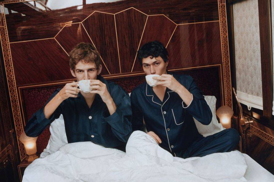 Zwei junge Männer in eleganten dunkelblauen Pyjamas sitzen in einem luxuriösen Bett mit dunklem Holzrahmen und Art-Déco-Details. Sie trinken stilvoll Tee aus feinen Porzellantassen und blicken ruhig in die Kamera. 
